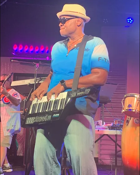 A musician plays a keytar on stage wearing a hat and sunglasses.