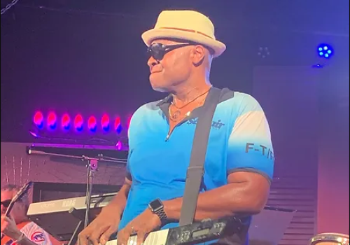 A musician plays a keytar on stage wearing a hat and sunglasses.